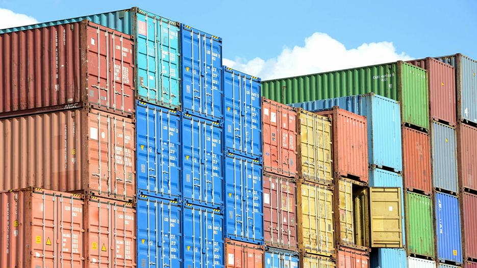 Stacks of colorful shipping containers, including blue, red, green, and yellow, arranged in rows under a bright blue sky.